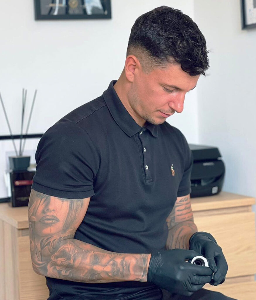 Tattoo Artist Andre Scharnagl aus Bremen in seinem Studio mit schwarzen Handschuhen, schwarzem Polo-Shirt und dekorativem Hintergrund